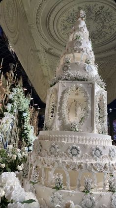 a large white cake with flowers on it and a clock at the top is surrounded by greenery