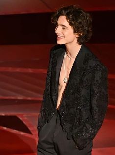 a man in a black suit and necklace standing next to a red wall with his shirt open