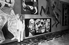 black and white photograph of man standing in front of wall with large paintings on it