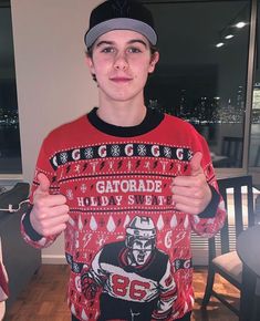 a young man wearing an ugly sweater and cap gives the thumbs up while standing in front of a window