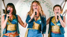 three women singing into microphones in front of a wall