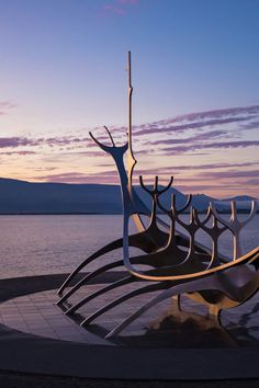 an artistic sculpture sits in front of the water at sunset with mountains in the background