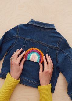 two hands are holding a crocheted rainbow brooch in front of a denim jacket