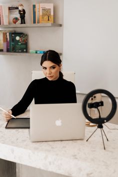 a woman sitting at a table with a laptop computer in front of her and a camera behind her