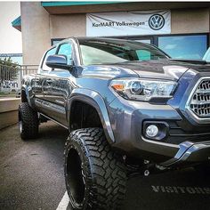 the front end of a gray truck parked in front of a building with large tires