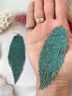 a hand holding two pairs of turquoise beaded earrings on top of a white table