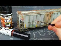 a hand is holding a pen and some paint next to a model train car on a table