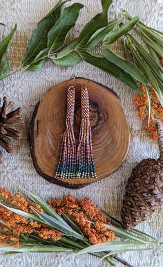 the beaded earrings are sitting on top of a piece of wood next to pine cones and leaves