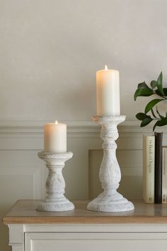 two white candles sitting on top of a table next to books and a potted plant