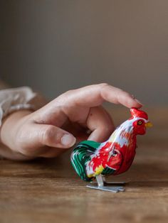 a small toy chicken sitting on top of a wooden table next to someone's hand