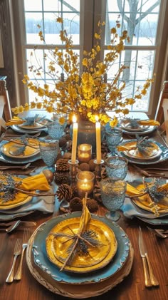 a table set with yellow and blue plates, silverware and candlesticks in front of a window