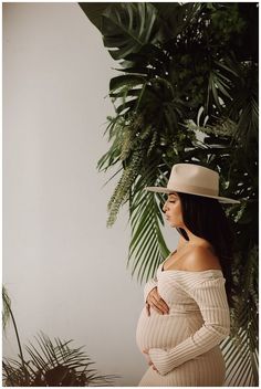 a pregnant woman wearing a hat and dress standing in front of a palm tree with her hands on her stomach