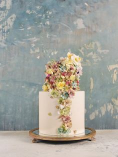 a white wedding cake with flowers on top and gold trimming around the bottom, sitting on a wooden stand in front of a blue wall