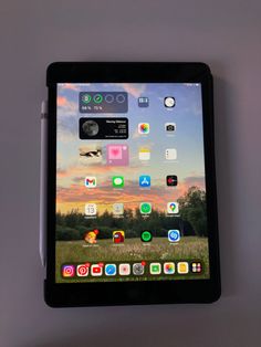 an ipad with icons on the screen is sitting in front of a gray wall and there are two pens next to it