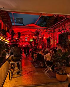 people are sitting in chairs at an outdoor bar with red lights on the ceiling and potted plants