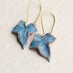 two blue leaves are hanging from gold earwires on a white surface, one is shaped like a leaf and the other is shaped like a flower