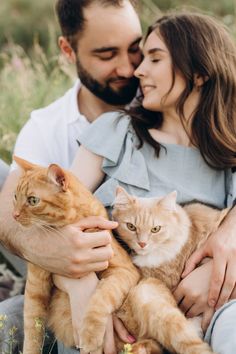 a man and woman holding two cats in their arms while sitting on the ground together