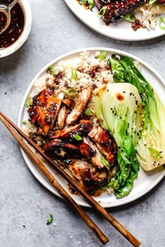 two plates filled with chicken and rice next to chopsticks on a gray surface