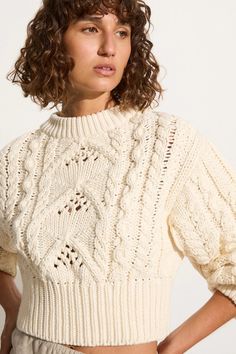 a woman with curly hair wearing a white knitted sweater and beige pants, standing in front of a white background