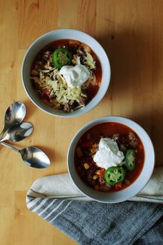 two bowls of chili with sour cream on top and spoons to the side sitting on a wooden table