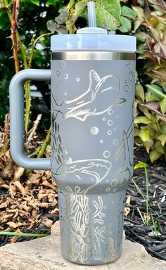 a stainless steel travel mug with a shark design on the lid and handle is sitting on a rock in front of some plants