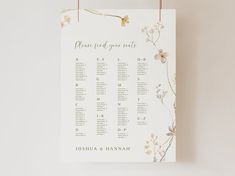 a seating chart hanging from a string on a white wall with flowers and leaves around it