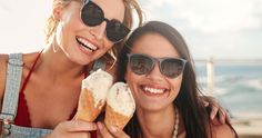 two beautiful women eating ice cream cones on the beach in front of the ocean and smiling