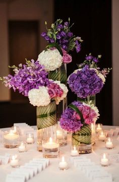 purple and white flowers are in vases on a table with lit candles around them