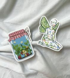 two stickers with fairy images on them sitting on a white cloth covered bed sheet