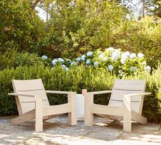 two wooden chairs sitting next to each other on a stone floor in front of bushes