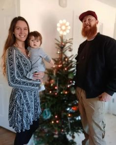 a man and woman standing in front of a christmas tree with a baby on their lap