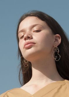 a woman with her eyes closed wearing large gold hoop earrings and a tan top against a blue sky