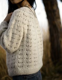 a woman standing next to a tree wearing a white sweater and blue jeans with her hands on her hips