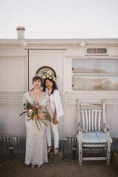 two women standing next to each other in front of a white trailer with flowers on it