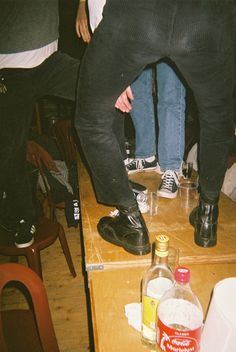 two people standing on top of a wooden table next to bottles and chairs in a room