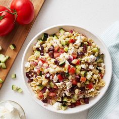 a white bowl filled with pasta salad next to tomatoes and cucumbers on a cutting board