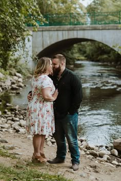 a man and woman standing next to each other in front of a river under a bridge