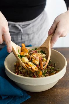 a person holding a wooden spoon over a bowl filled with vegetables and grains, on top of a table