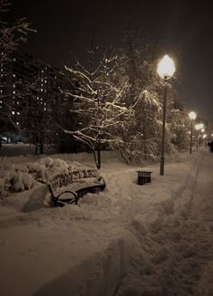 a park bench covered in snow next to a street light at night with people walking on the sidewalk