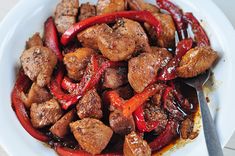 a white plate topped with meat and red peppers next to a fork on top of a table