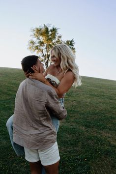 a man and woman hug each other in the middle of a grassy field with a tree behind them