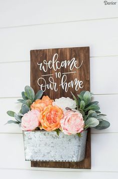 a welcome sign hanging on the side of a building with flowers in it and greenery