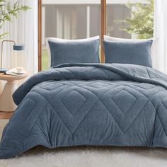 a bed with blue comforter and pillows in front of a window, next to a potted plant