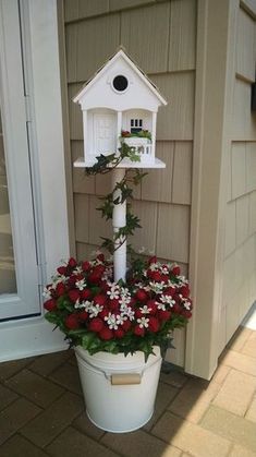 a white birdhouse sitting on top of a bucket filled with red and white flowers