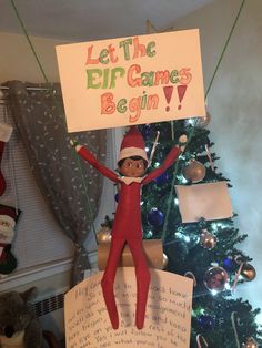 an elf holding up a sign in front of a christmas tree