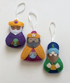 three felt ornaments are hanging from strings on a white surface, one has an ornament in the shape of two wise men