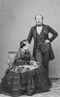 an old black and white photo of two people in formal dress sitting on a chair