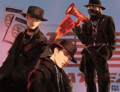 three men in hats and black clothing stand next to each other, one holding an orange bat