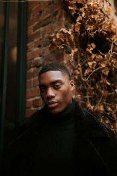 a man standing in front of a brick building wearing a black turtle neck sweater and coat