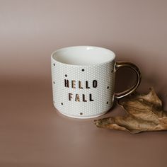 a coffee mug with the words hello fall written on it next to a fallen leaf